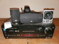 Panasonic AV control receiver with four black JBL speakers of various sizes arranged on top of it, showing front view of receiver with labels and JBL logos on speakers.