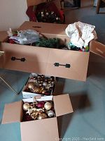 Boxes containing assorted Christmas balls, garland, artificial flowers, and a tree skirt, showing overall lot contents.