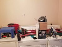 Full lot view on washer and dryer including multiple brooms, Black & Decker Dustbuster, steam iron, Vileda mop bucket, blue plastic stool and small cleaning tools in red bucket.