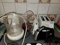 Four electric kitchen appliances grouped together on a stove, including a food processor, two hand blenders with whisk attachments, and a hand mixer.