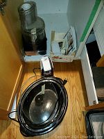 Photo showing the set of kitchen appliances including food processor with accessories inside a cabinet, and can opener on the floor with multiple black flat cooking trays, an oval black pan and silver oven guard rack.