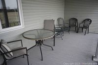 Outdoor patio furniture on a wooden deck, showing round metal and glass table with two cushioned metal chairs and other seating against siding wall.