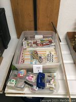 Plastic bin full of assorted crafting papers, stickers, and embellishments with blue T-square and small tools in foreground
