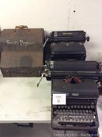 Three vintage manual typewriters displayed side by side: Smith Premier with carrying case, Royal, and Remington models.