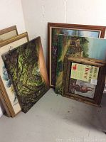 Framed signed art pieces stacked against a wall showing visible details of the artwork and frames including Pontiac print with crack in glass.