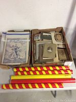 Two boxes on a table; box one contains vintage photos and ephemera, box two has sheet music with visible covers, and red/yellow striped wooden rods lying in front