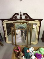 Four mirrors stacked together: cream ornate mirror, three-panel brass vanity mirror on top, large wooden-frame mirror with decorative top behind them.