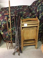 Photo showing four wooden folding chairs stacked together, old golf clubs with metal club heads and wooden shafts, and two garden rakes with wooden handles and metal tines.