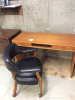 Full view of the wood two-drawer desk with central open shelf and the black upholstered chair with wooden legs and caster wheels.