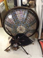 Underside view of large stained glass shade resting on the floor with chandelier frame and mesh panels beneath
