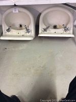 Two older single basin off-white sinks with chrome faucets, visible signs of use, mounted on the wall or displayed together in a storage or sale setting.