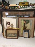 Multiple framed art pieces stacked and leaning, showing variety including a quilt square and lithographs.