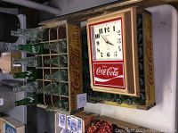 Photo showing one wooden Coca-Cola crate filled with vintage empty glass bottles, and a Hanover Coca-Cola clock placed on top of another crate.