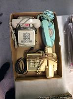Three vintage telephones in a cardboard box showing top side with handset and keypads visible.