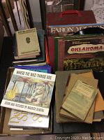 Top view of stacks of old books and vinyl records in sleeves. Visible books include 'Where the Wild Things Are' and a 'Roy Blakeney First Series History' book. Several vinyl record sleeves including Faron Young Greatest Hits and Oklahoma soundtrack.