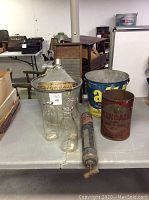 Full view of lot with Kendall oil can, All bucket, three milk bottles, metal funnel, metal bucket, duster on table. Visible vintage typewriter and washboard in background.