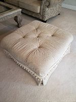 Square beige fabric ottoman with tufted top cushion next to a beige patterned sofa and decorative wood coffee table.