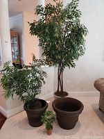 Overall photo showing the three artificial trees/plants and two planters grouped on carpeted floor near corner walls in living space.