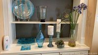 Shelf display showing tall blue bubbled glass vases and trays, glass plate with metal stand, ceramic candle holders and small succulent in a pot.
