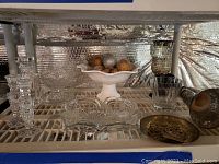 Wide view showing various cut glass candlestick holders, candy bowls, mercury glass votive holder, white ceramic pedestal bowl with glittery artificial fruit, and brass-tone dish on display shelf.