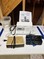 Panasonic fax machine, operating instructions, Capello alarm clock in box, small bin of air freshener wall outlets, cords visible on table under stairs.