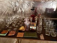 Wide view showing multiple clear glass wine glasses, cocktail glasses, pitchers, travel cups, salt and pepper shakers and drink coasters on a table with reflective silver backdrop.