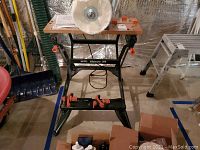 Front view of the Black & Decker Workmate 200 workbench with wooden top and two black and orange clamps attached to the metal front bar, standing on a concrete floor with tools and boxes nearby.