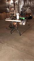 Full view of plastic folding table in basement setting with other belongings around, showing table surface and black folding legs.