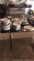 Wide view showing all stacked China Pearl plates and cups, Gibson dishes on folding table in basement