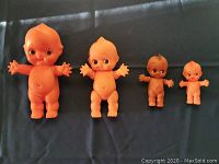 All four Kewpie dolls lined up by size on black background: 9 inch and 8 inch orange rubber squeaky dolls, 5 inch black rubber doll, 4 inch plastic doll.