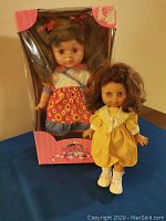 Two soft plastic dolls displayed. One doll is posed standing outside the box wearing a yellow dress and white shoes. The other doll is in original packaging wearing a red-orange floral dress with apron pattern and blue skirt.