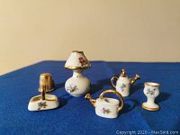 Five pieces of Limoges porcelain miniatures displayed on blue cloth with a thimble included for scale reference, showing a lamp, two watering cans, a cup, and a basket-like item, all with floral patterns and gold accenting.