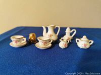 Angle showing the entire miniature tea set on blue cloth background, including two cups with saucers, teapot (no lid), sugar bowl with lid, creamer, small pitcher, and gold thimble used for size comparison.