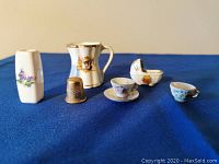 Six miniatures displayed on blue fabric: a small rectangular vase with purple flower design, a metal thimble (for size comparison), a Queen Elizabeth II cup, a white bassinet figurine with flower decal, two mini teacups (one on saucer).