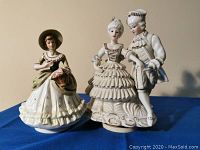 Two musical wind-up porcelain figurines displayed side by side on a blue cloth background, showing detailed dress and coloration.