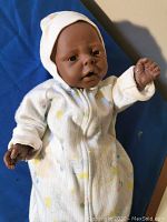 Full view of the Jesmar African American newborn doll in white patterned sleeper and white hat, laying on blue background.