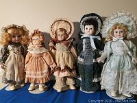 Five porcelain dolls standing side by side in vintage-style dresses with bonnets and aprons on display on blue cloth background.