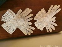 Two pairs of handmade white crochet gloves laid flat on a wooden surface showing their full length and scalloped edges.