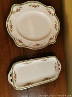 Both platters shown on wood surface. Oval platter has scalloped edges and decorative floral and green border pattern. Rectangular platter has cutout handles with matching floral design.