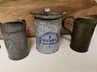 Three metal measuring cups including a speckled gray 1 quart cup with blue lettering, one plain metal 1 pint cup with patina, and one brass-colored 1 pint cup marked General Steel Wares.