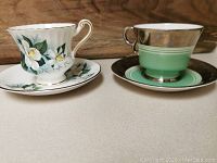 Two vintage tea cup and saucer sets displayed side by side on a flat surface with a wood background, showing floral and green/gold designs.