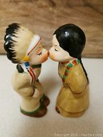 Front side of the kissing salt and pepper shakers showing the male figure with feathered headdress and female figure with black hair in traditional attire.