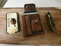 Three items displayed on wood: a vintage Panther cigarette tin, a brown leather belt pouch with stitched pockets and flap, and an old pocket knife with a worn handle.