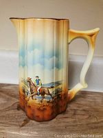 Pitcher showing handpainted scene of man riding horse in rural landscape with blue sky and clouds background.