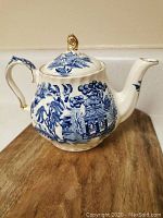 Front and side view of white ceramic tea pot decorated with blue painted Asian pagoda scene and gold accents on handle and lid knob.