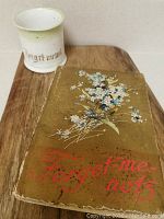 Photo showing the antique Forget Me Nots book with floral cover and the Forget Me Not cup on a wooden surface.