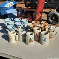 Group photo of assorted steins and goblets on a white table with outdoor background showing all items in the lot