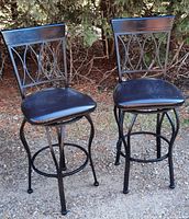 Front view of two black metal bar chairs with lattice backrests and padded black seats on gravel ground with shrubs in background.