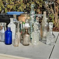 All vintage glass bottles arranged on white surface outdoors with natural light. Variety of shapes, sizes, and colors visible.