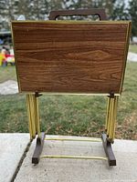 View of the front side of one TV tray in folded position showing wood grain laminate top with brass edge trim and dark brown plastic feet supporting brass frame.